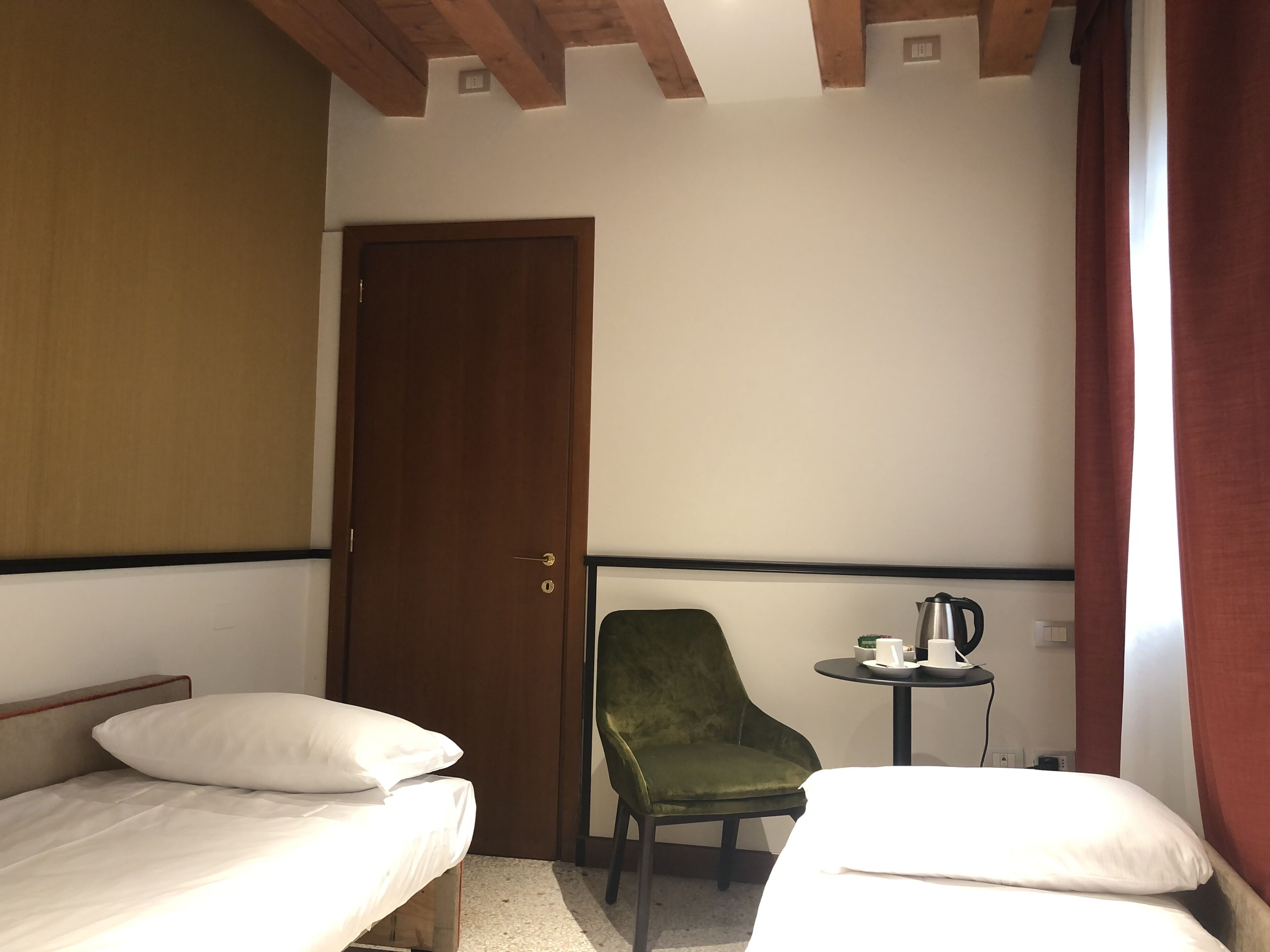 A small, simple room with two single beds, a green chair, a small table with a kettle, and a door.