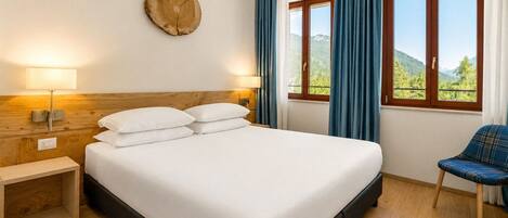 A hotel room with a bed, bedside lamps, a wooden headboard, a chair, a window with curtains, and a wall-mounted clock.