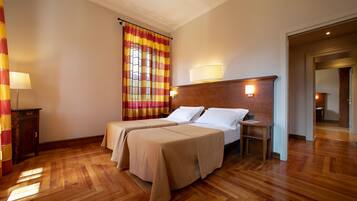A hotel room with two beds, wooden flooring, a window with striped curtains, a bedside table with a lamp, and a door leading to another room.