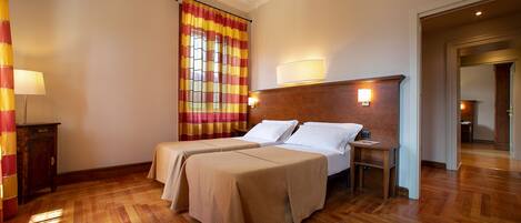 Une chambre d’hôtel avec deux lits, un plancher en bois, une fenêtre avec des rideaux à rayures, une table de chevet avec une lampe et une porte donnant sur une autre pièce.