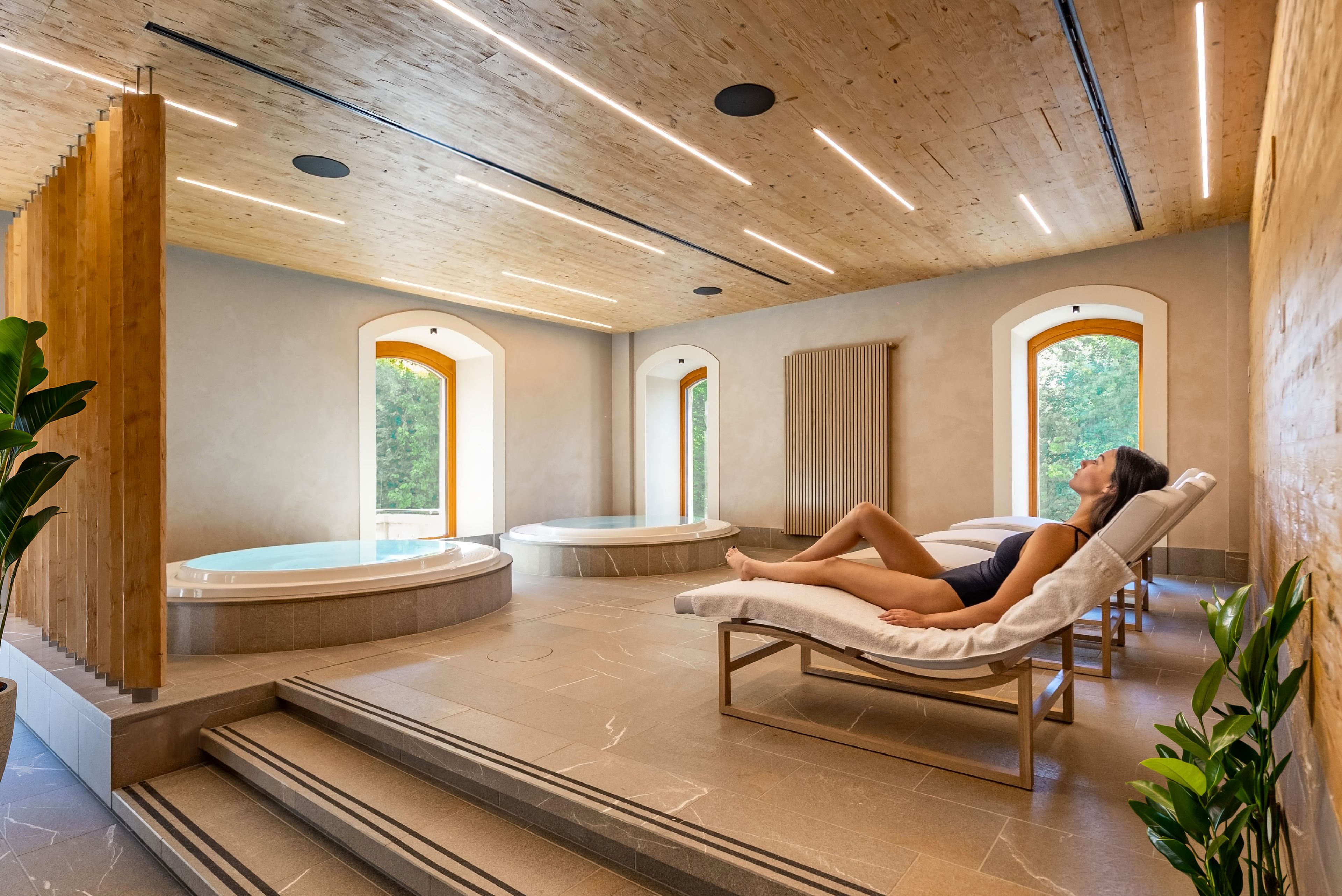 Ein Wellnessraum mit einer Person, die auf einer Liege entspannt, einer Whirlwanne und Blick auf Grün durch Bogenfenster.