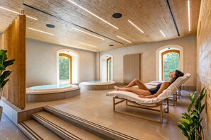 Un cuarto de spa con una persona relajándose en una tumbona, un jacuzzi y vistas a la vegetación a través de ventanales arqueados.