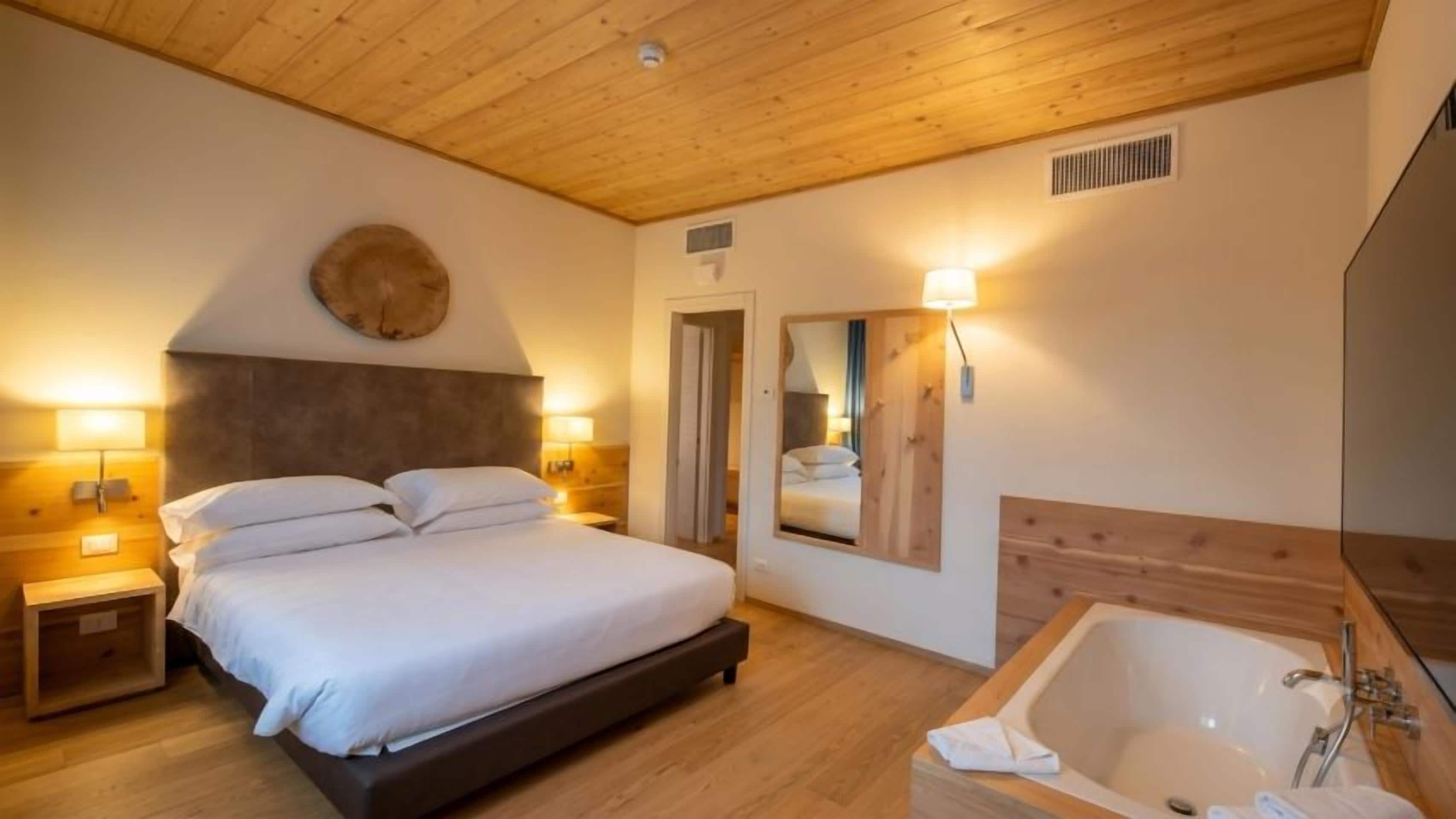 A bedroom with a bed, bedside lamps, a large mirror, a wooden headboard, and a bathtub.