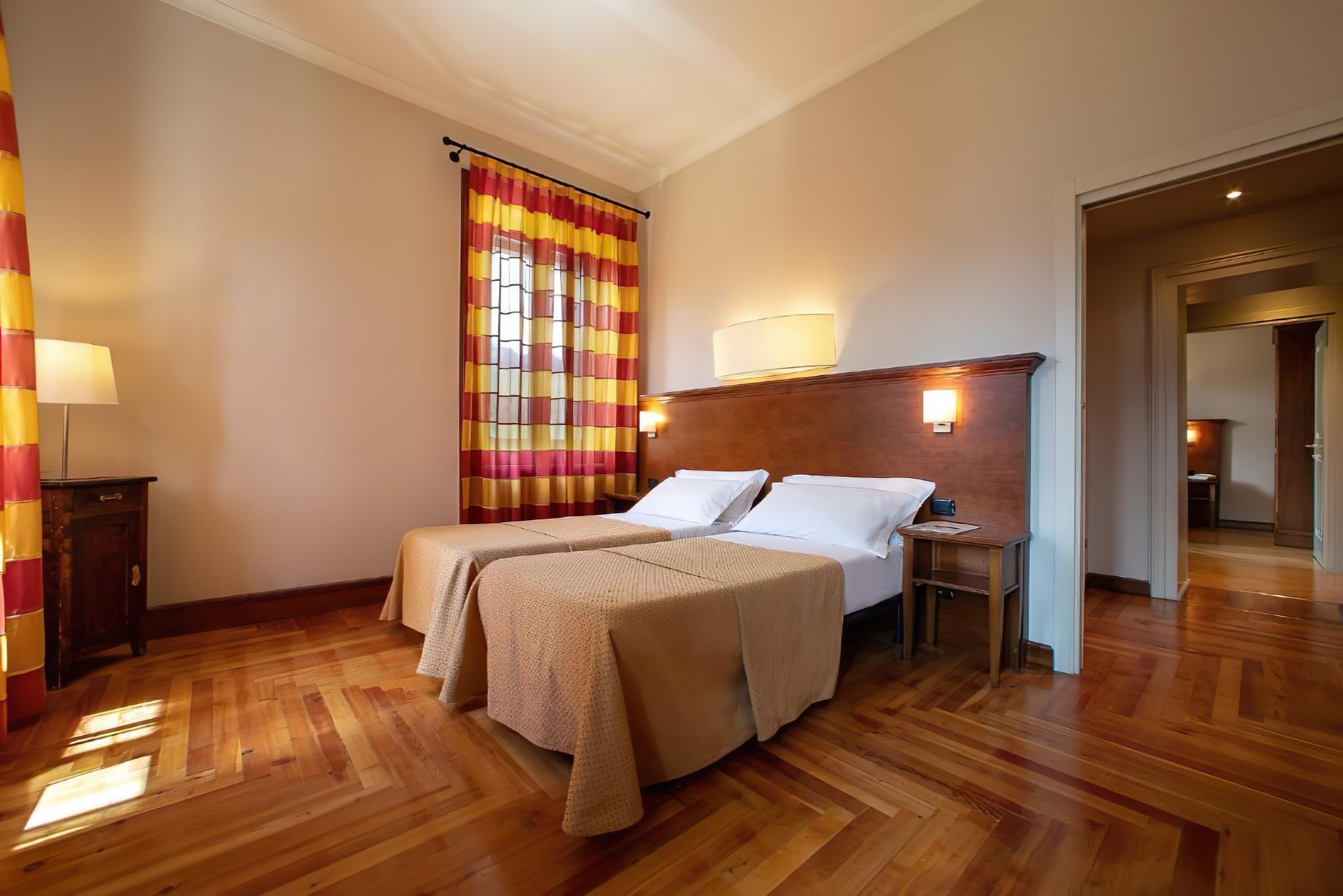 A hotel room with a double bed, a nightstand, a lamp, a window with curtains, and a wooden floor.