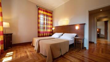 A hotel room with a double bed, a nightstand, a lamp, a window with curtains, and a wooden floor.