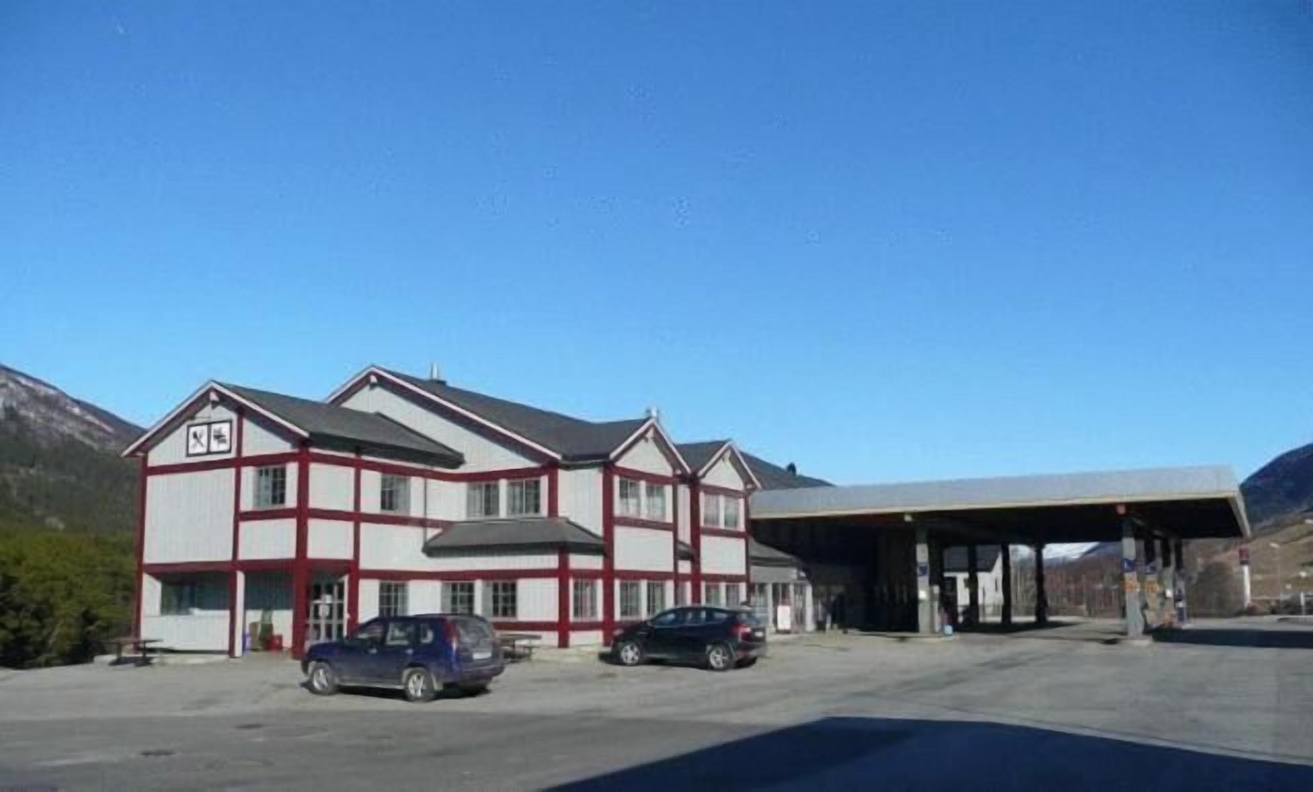 Un edificio de dos pisos con fachada roja y blanca, zona de estacionamiento techada y montañas al fondo.