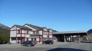 Un edificio de dos pisos con fachada roja y blanca, zona de estacionamiento techada y montañas al fondo.