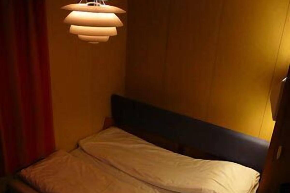 A small, dimly lit room with a single bed, a wooden headboard, and a hanging light fixture.