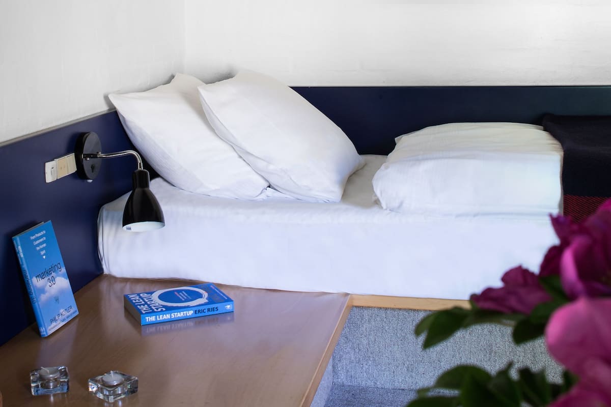 A neatly made bed with white linens, a bedside lamp, a book titled "The Lean Startup Eric Ries", and a small ashtray on a wooden nightstand.