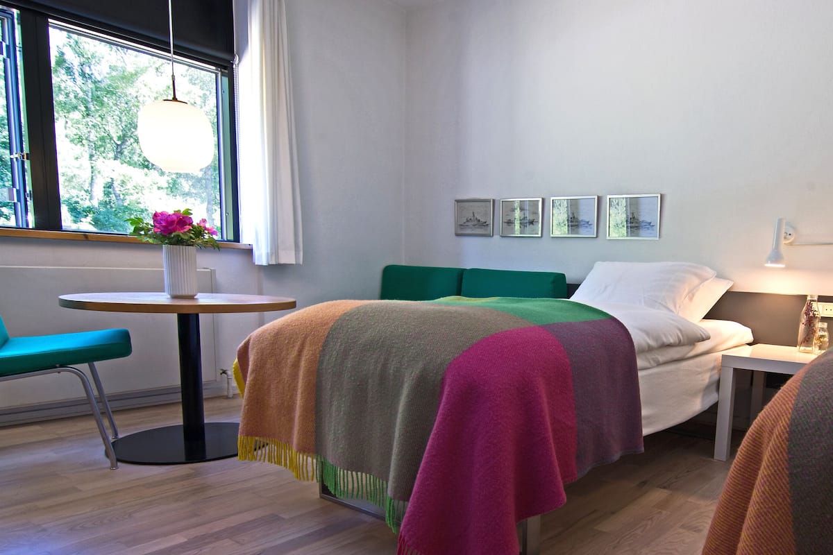 A hotel room with two beds, a small table, a chair, and a window with a view of trees.