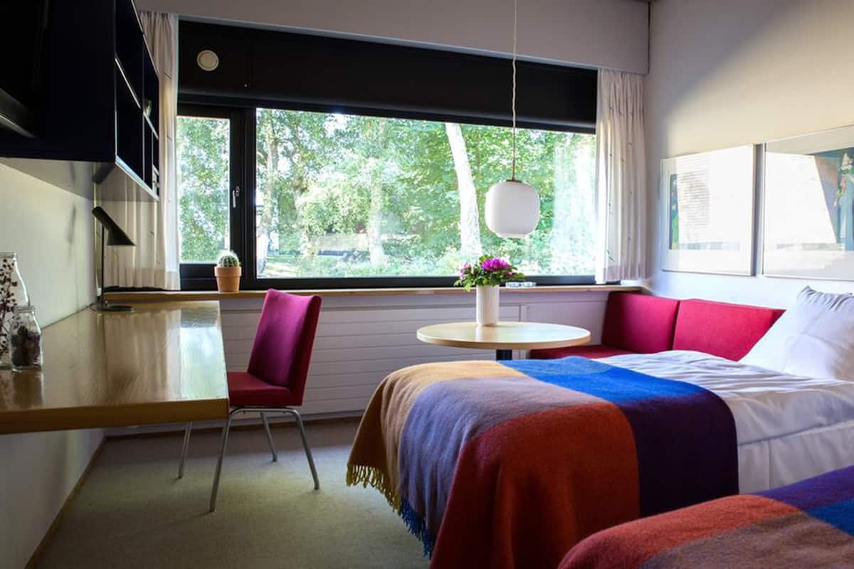 A modern hotel room with a large bed, a red sofa, a small table, and a window with a view of greenery.