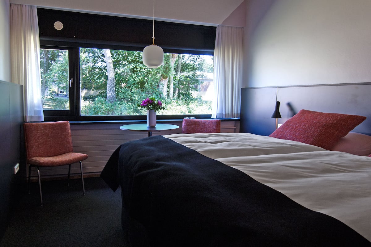 A hotel room with a bed, two red pillows, a chair, a small round table with a vase of flowers, and a window with a view of trees.