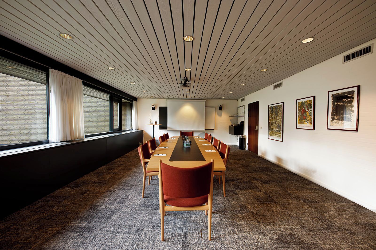 A conference room with a long table, red chairs, a projector screen, and framed artwork on the walls.