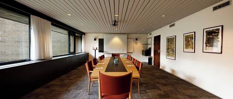 A conference room with a long table, red chairs, a projector screen, and framed artwork on the walls.