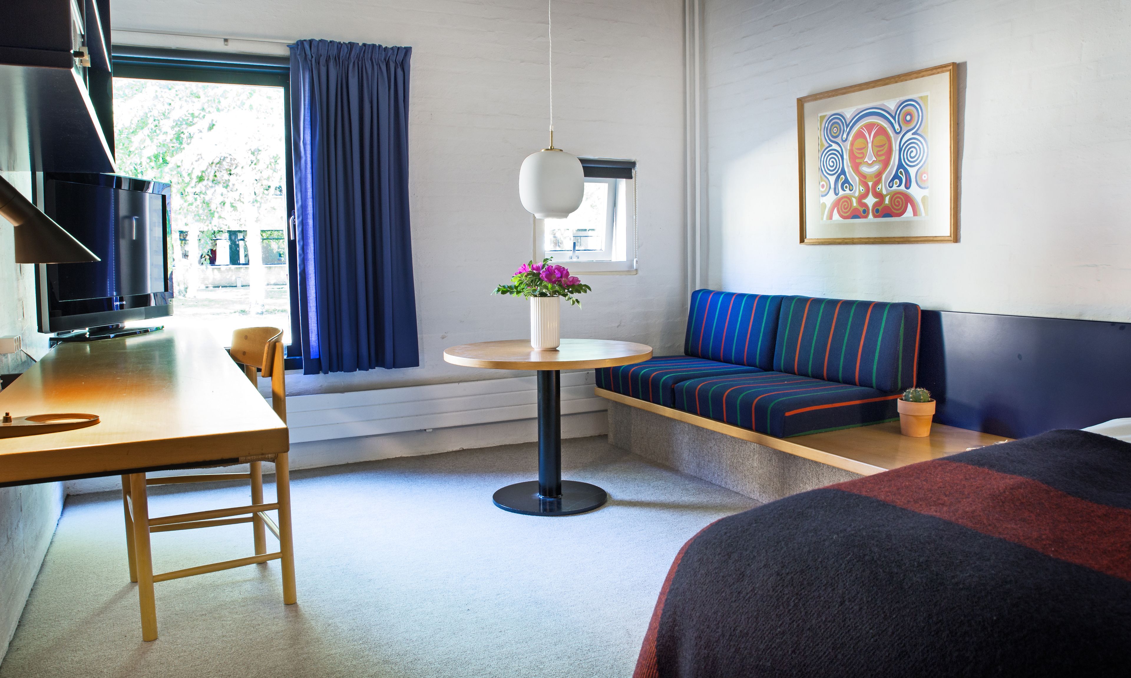 Et hotelværelse med en seng, en sofa, et lille bord med en vase blomster, et fjernsyn og et vindue med gardiner.
