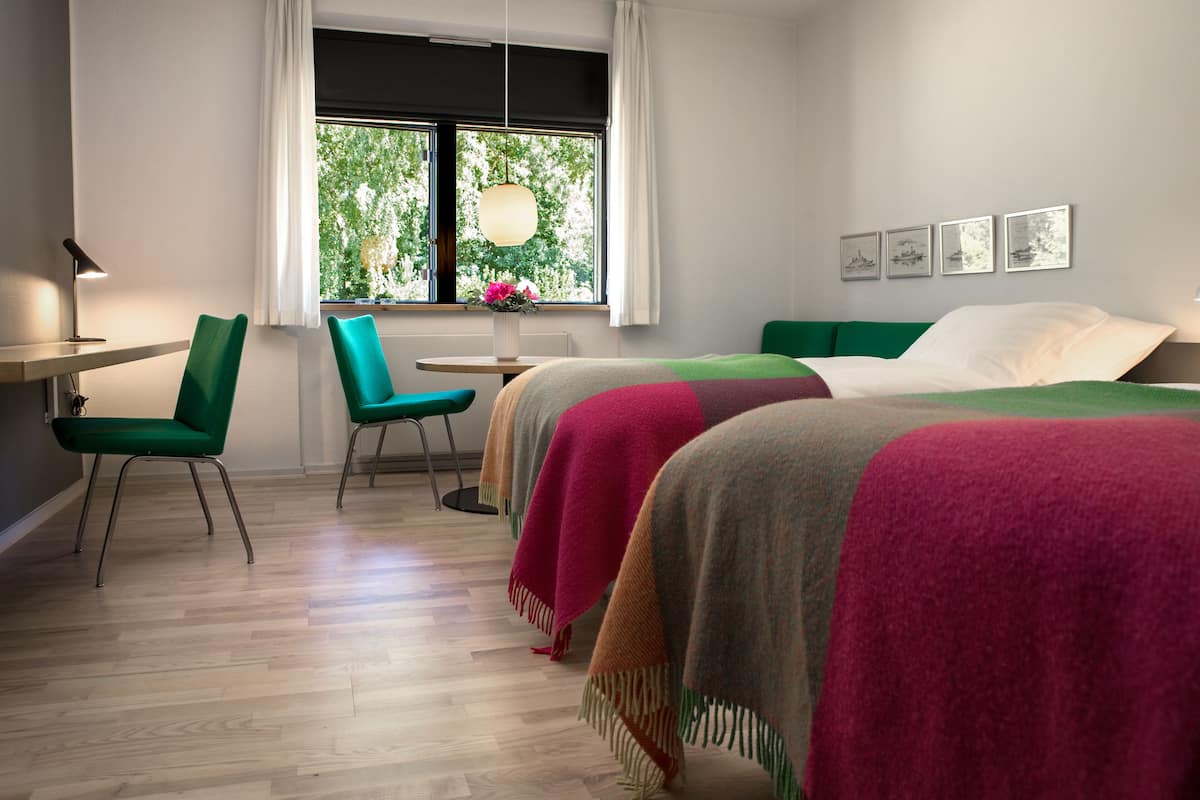 A hotel room with a wooden floor, two green chairs, a small table with a vase of flowers, a bed with colorful blankets, a desk with a lamp, and a window with a view of greenery.