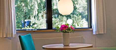 A room with a window, a teal chair, a small table, and a vase with flowers.