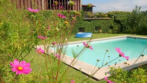 Uma piscina cercada por vegetação exuberante e flores cor-de-rosa, com uma casa ao fundo.