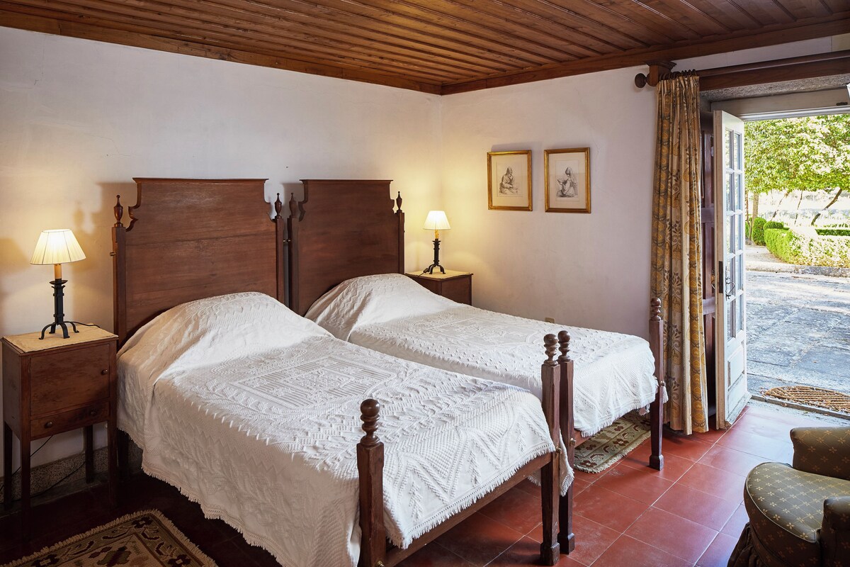 Una habitación con dos camas de madera, cada una con una colcha blanca y una mesita de noche con una lámpara. Hay dos cuadros enmarcados en la pared y una ventana con cortinas.