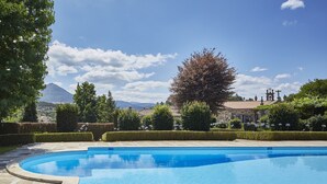 Una piscina con solárium de piedra, rodeada de setos y árboles cuidados, con una montaña y un edificio al fondo.