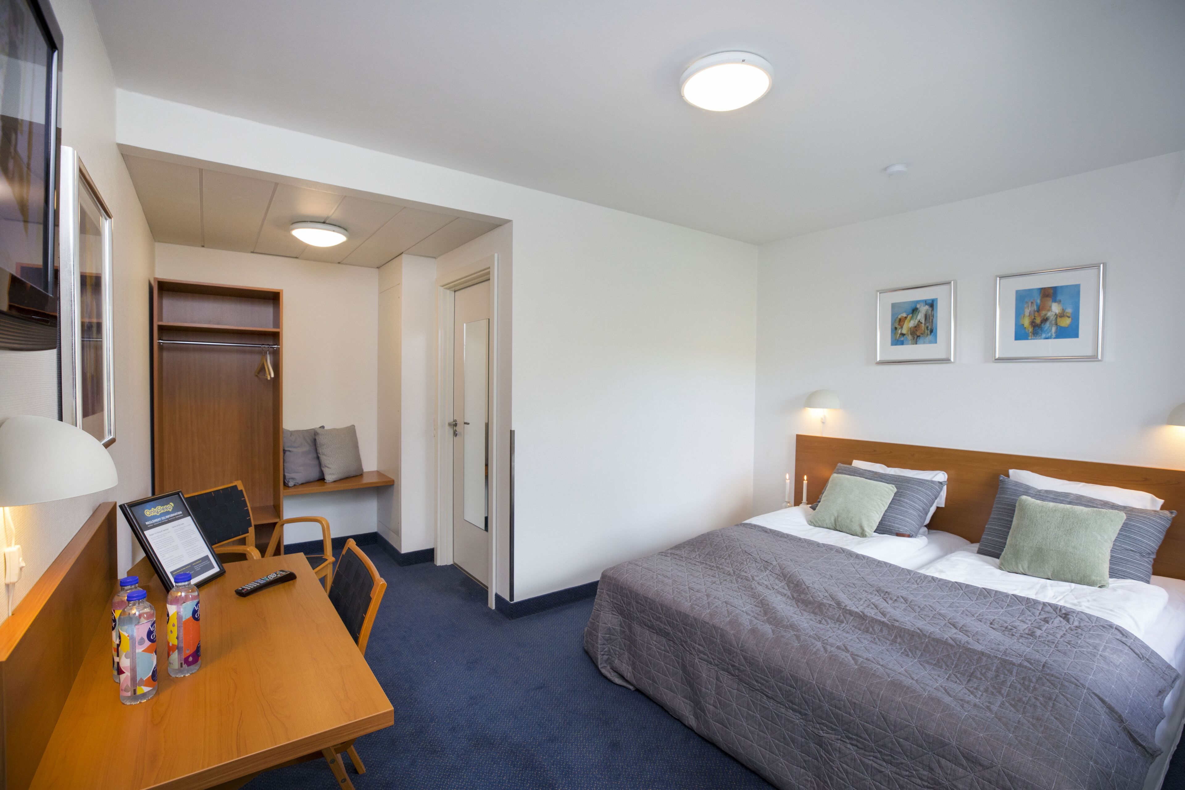 Camera d'albergo con un letto, una scrivania con un computer e due bottiglie, una sedia e una porta che conduce a un'altra stanza.