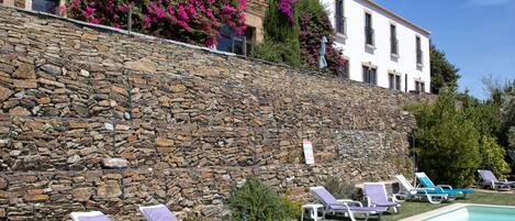 Zona de piscina con sillones de descanso y un edificio de piedra con fachada blanca.