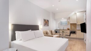 Habitación de hotel moderna con una cama grande, una pequeña mesa redonda, un sofá, un televisor y una kitchenette al fondo.