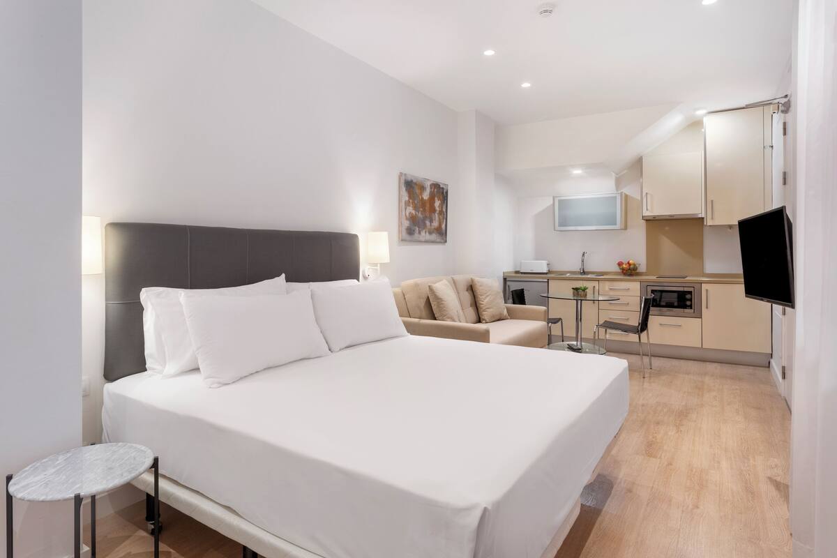 Habitación de hotel moderna con una cama grande, una pequeña mesa redonda, un sofá, un televisor y una kitchenette al fondo.