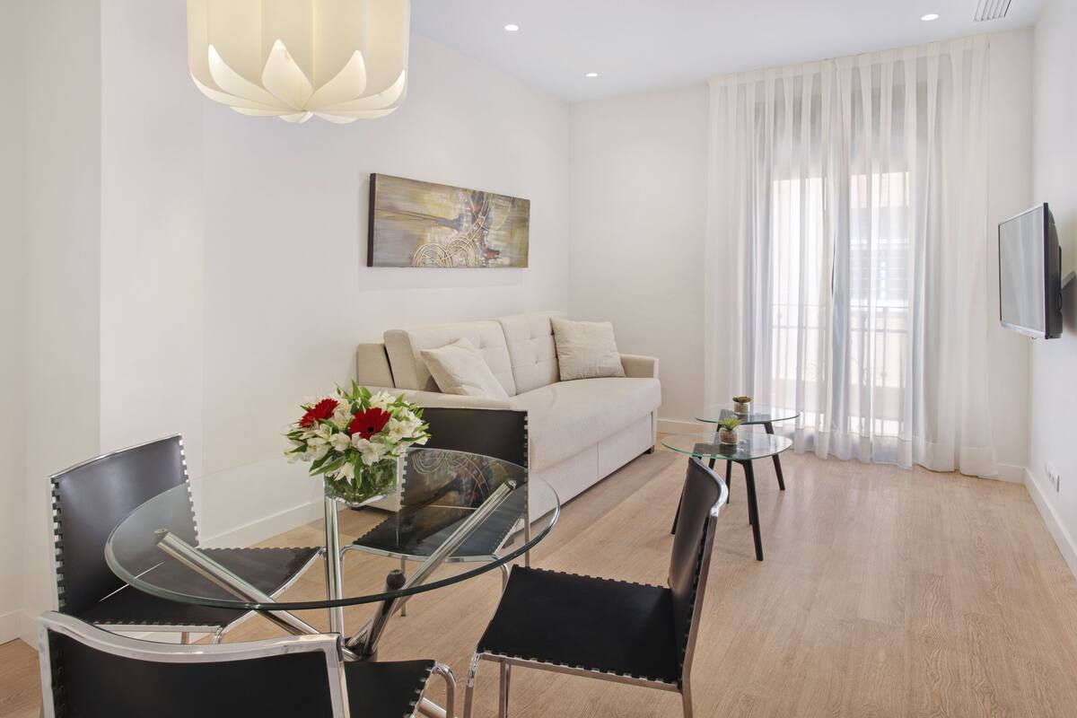 Una sala de estar moderna con una mesa de comedor de vidrio, sillas negras, un sofá color beige y un ventanal con cortinas translúcidas.