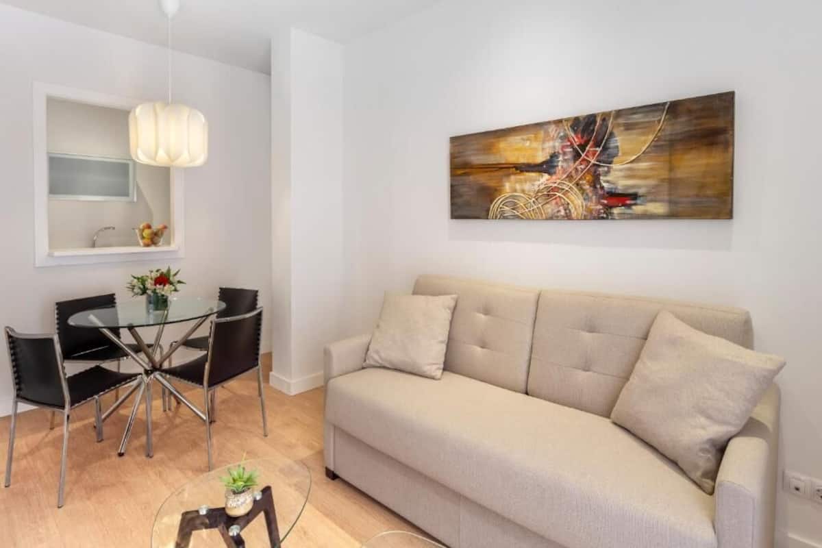 A modern living room with a beige sofa, a glass dining table with chairs, a pendant light, and a large abstract painting on the wall.