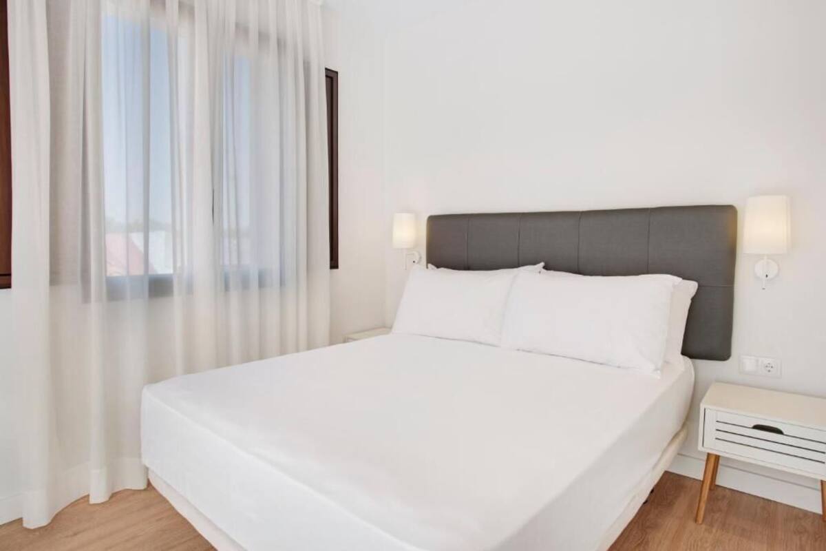Una cama bien hecha con sábanas blancas en una habitación con suelo de madera, paredes blancas y una ventana con cortinas de gasa.
