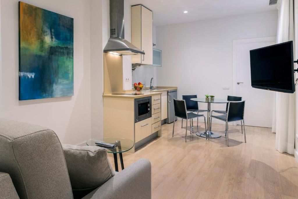 Una sala moderna con sofá, una mesa de centro de vidrio, un área de comedor con mesa y sillas, una cocina con estufa y armarios, y un televisor de pantalla plana montado en la pared.
