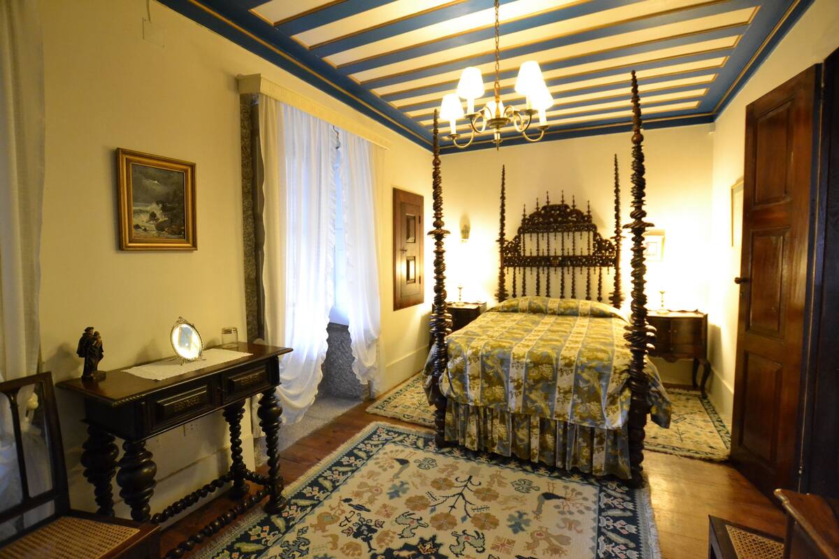 A four-poster bed with a floral bedspread, a wooden nightstand with a lamp, and a desk with a mirror and a small statue.