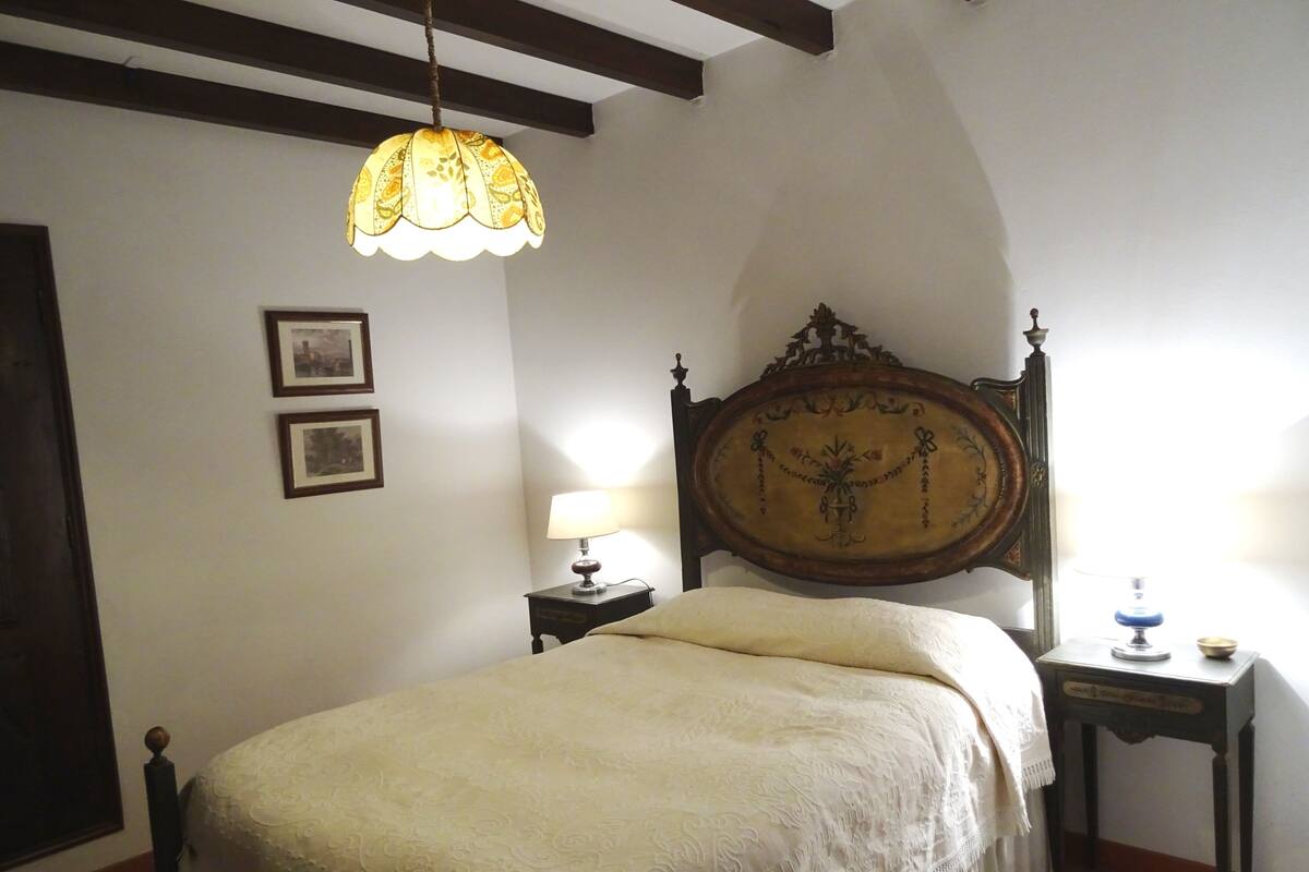 A bedroom with a large bed, a wooden headboard, a hanging lamp, a bedside table, and two framed pictures on the wall.