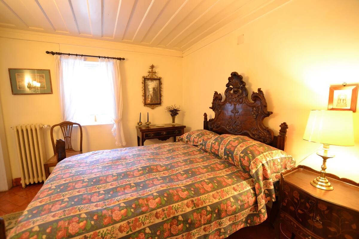 A bedroom with a floral bedspread, a wooden headboard, a bedside table with a lamp, a chair, and a radiator.