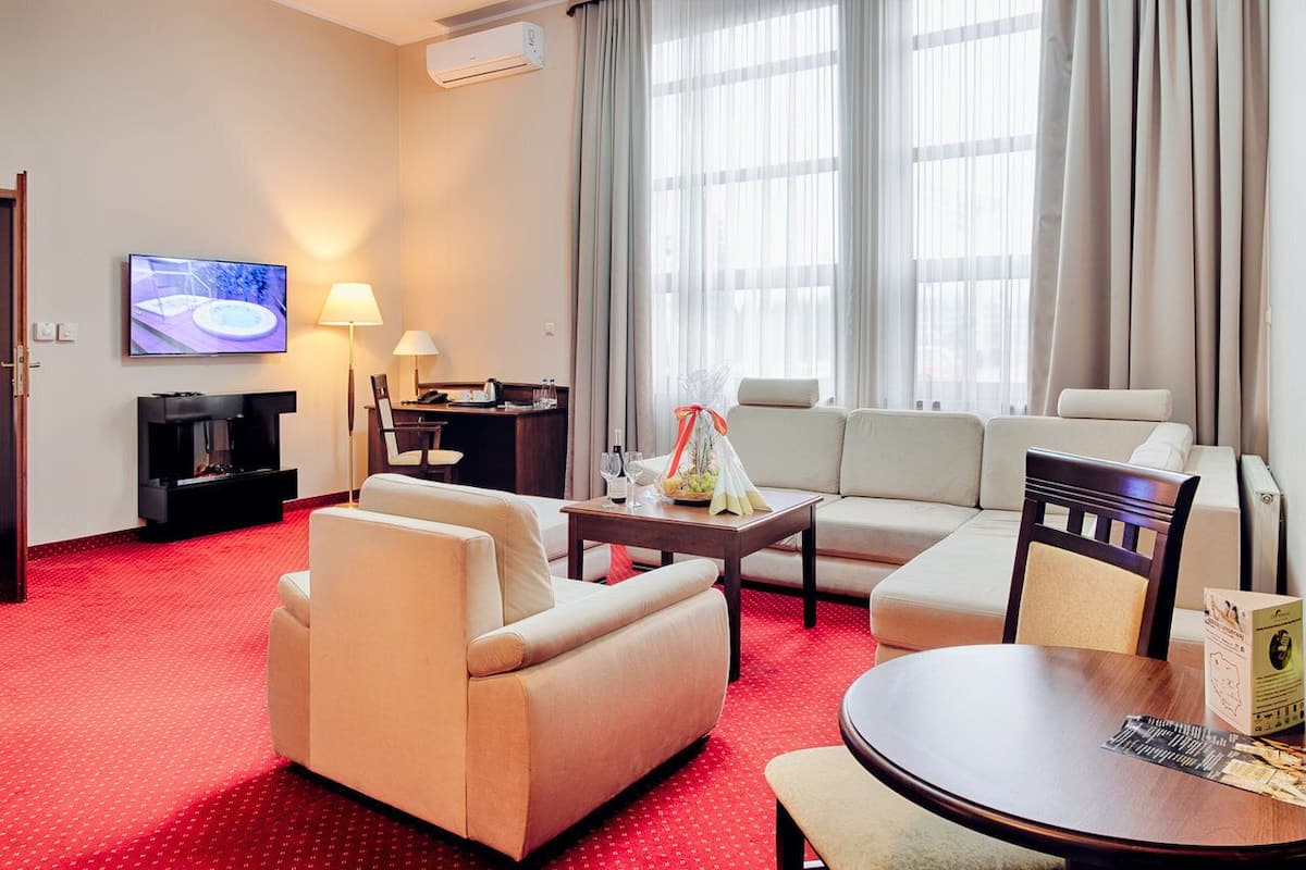 A hotel room with a red carpet, a sofa set, a TV, a desk with a lamp, and a large window with curtains.