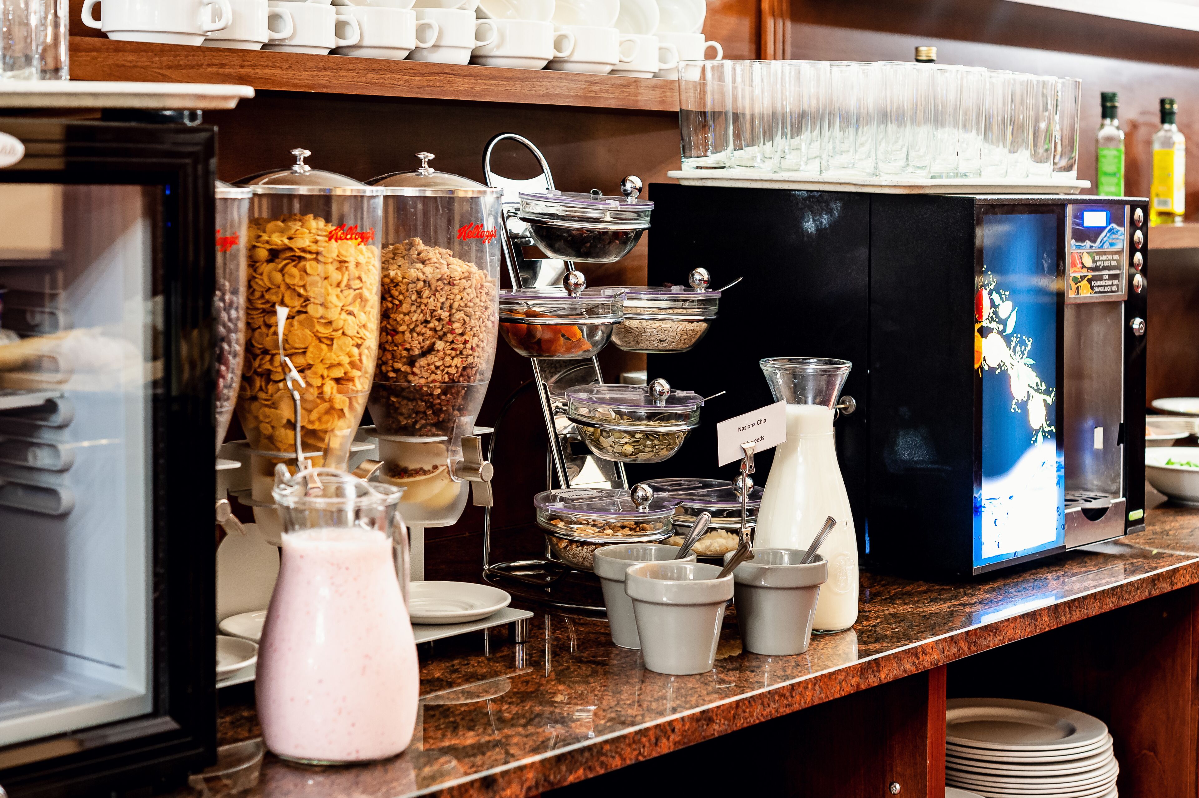 Zona de bufé con diferentes dispensadores de comida y bebida, incluyendo cereales, granola y leche.