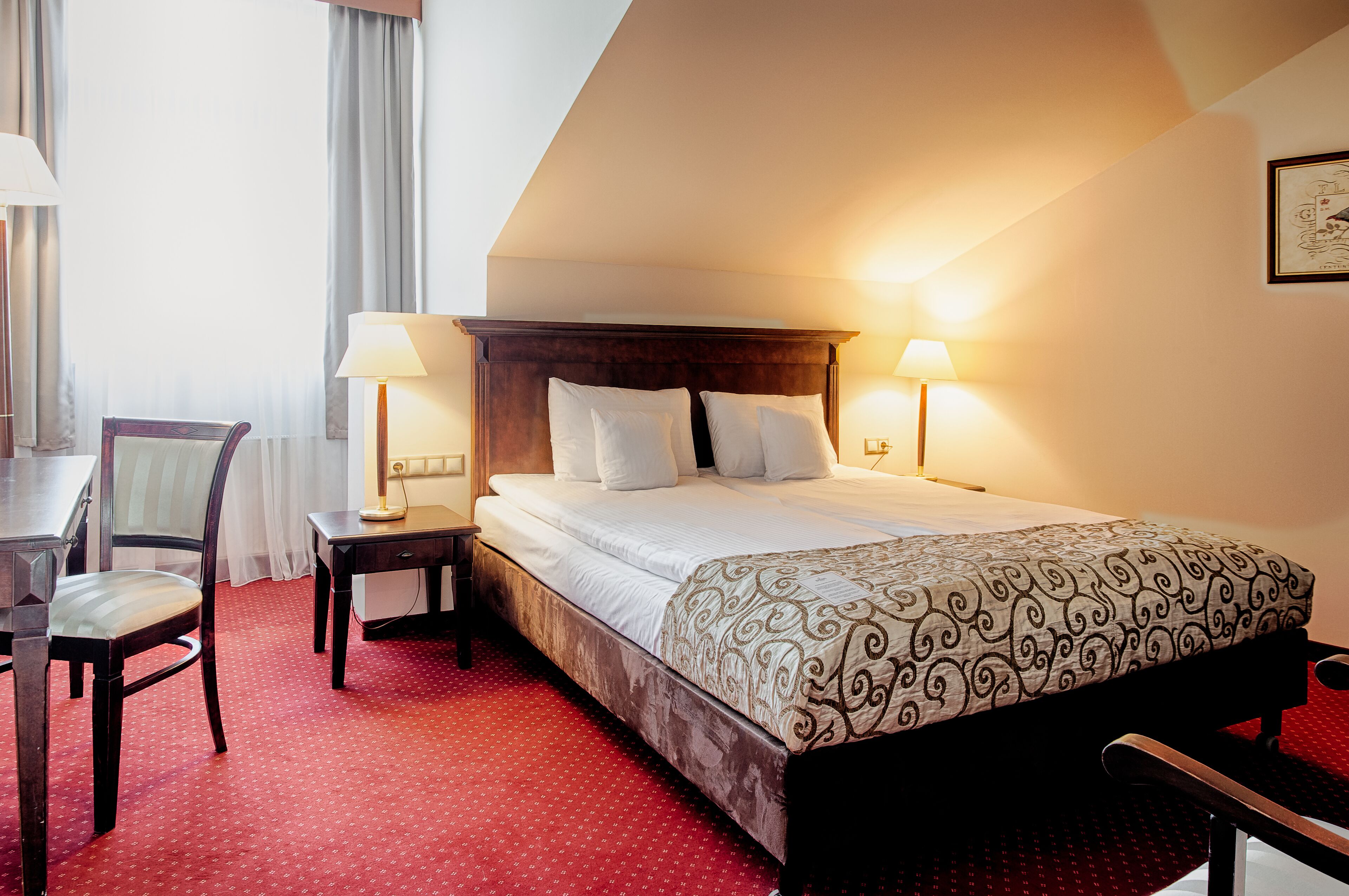 A hotel room with a bed, two bedside lamps, a desk with a chair, and a red carpeted floor.