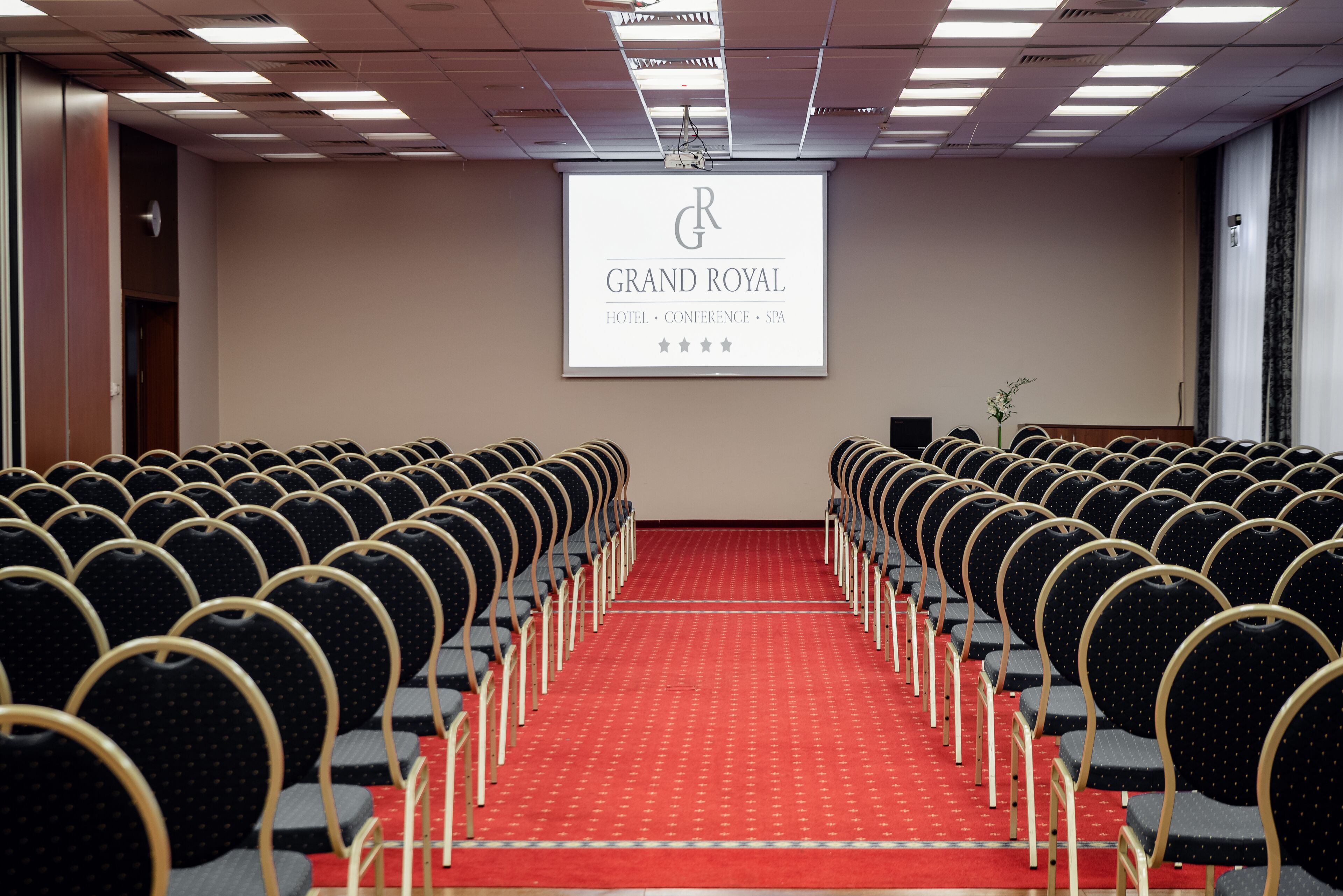 A conference room with rows of chairs and a red carpet, a projector screen displaying "GRAND ROYAL HOTEL - CONFERENCE - SPA" with a star rating, and a small plant on the right.