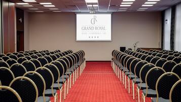 Uma sala de conferências com fileiras de cadeiras e um tapete vermelho, uma tela de projetor exibindo "GRAND ROYAL HOTEL - CONFERÊNCIA - SPA" com classificação por estrelas, e uma pequena planta à direita.