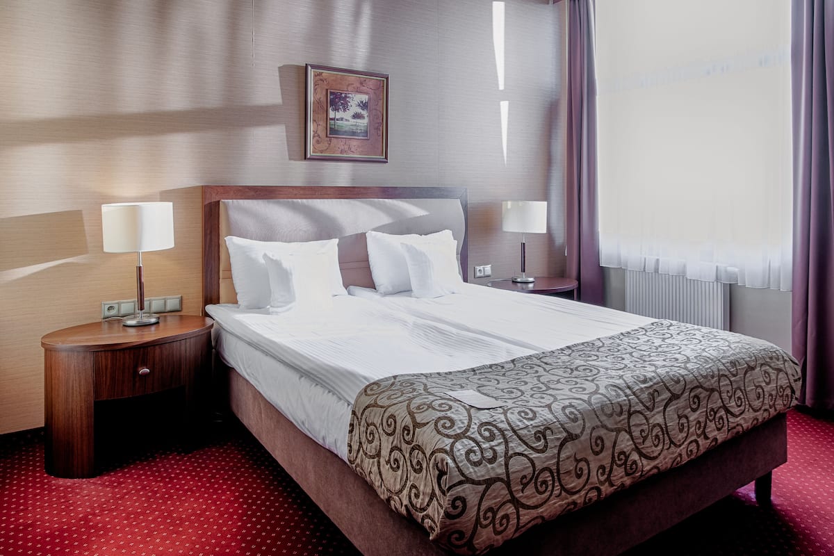A hotel room with a neatly made bed, two bedside tables with lamps, a framed picture on the wall, and a window with curtains.