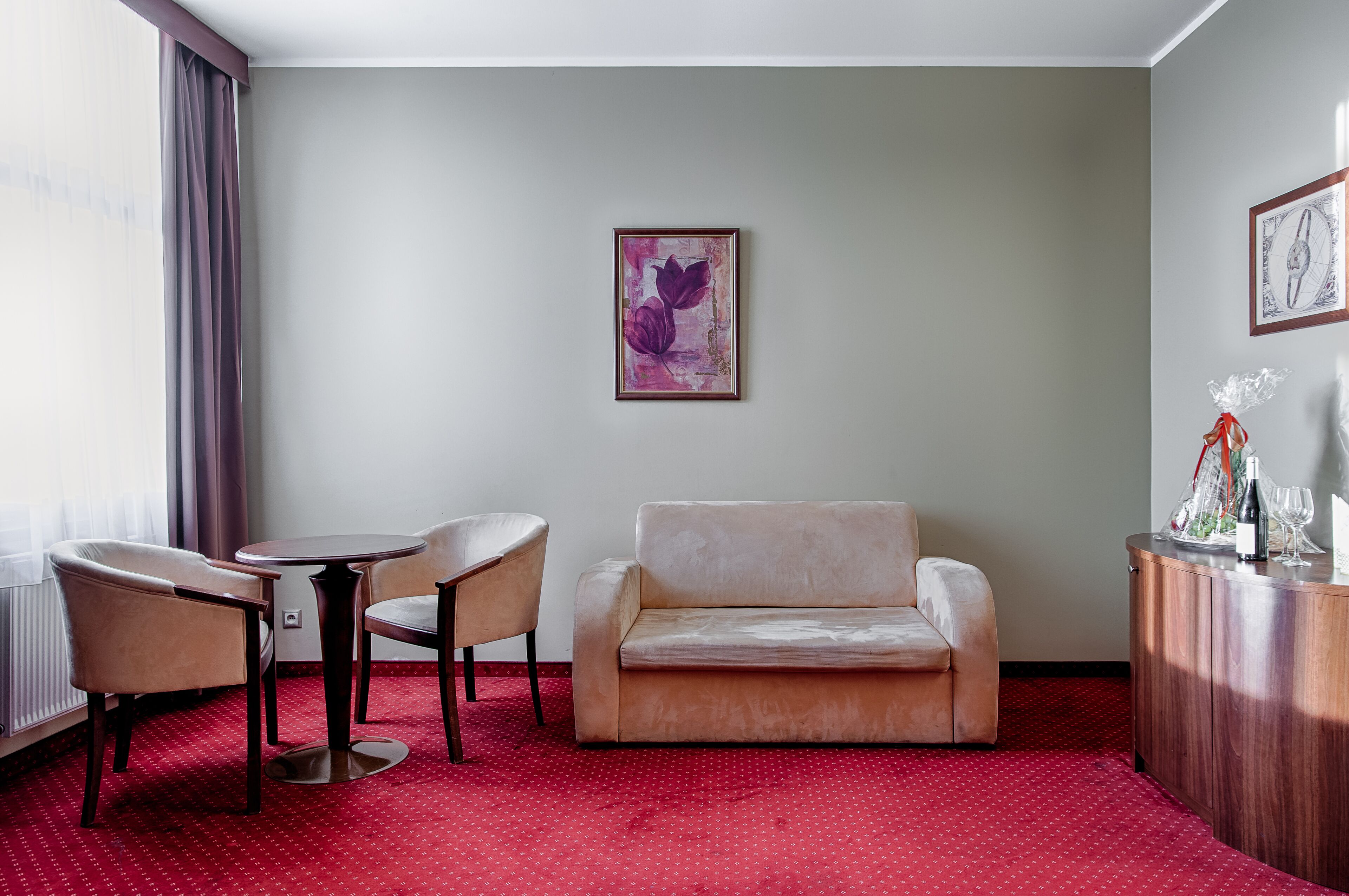 Habitación de hotel con alfombra roja, un sofá, dos sillones, una mesa redonda pequeña y un aparador con una botella y vasos.