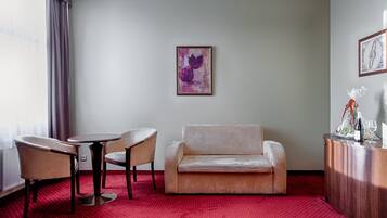 Habitación de hotel con alfombra roja, un sofá, dos sillones, una mesa redonda pequeña y un aparador con una botella y vasos.