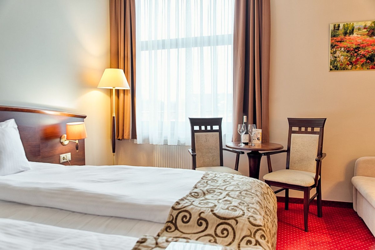 A hotel room with a bed, two chairs, a small table with a wine glass, and a painting on the wall.