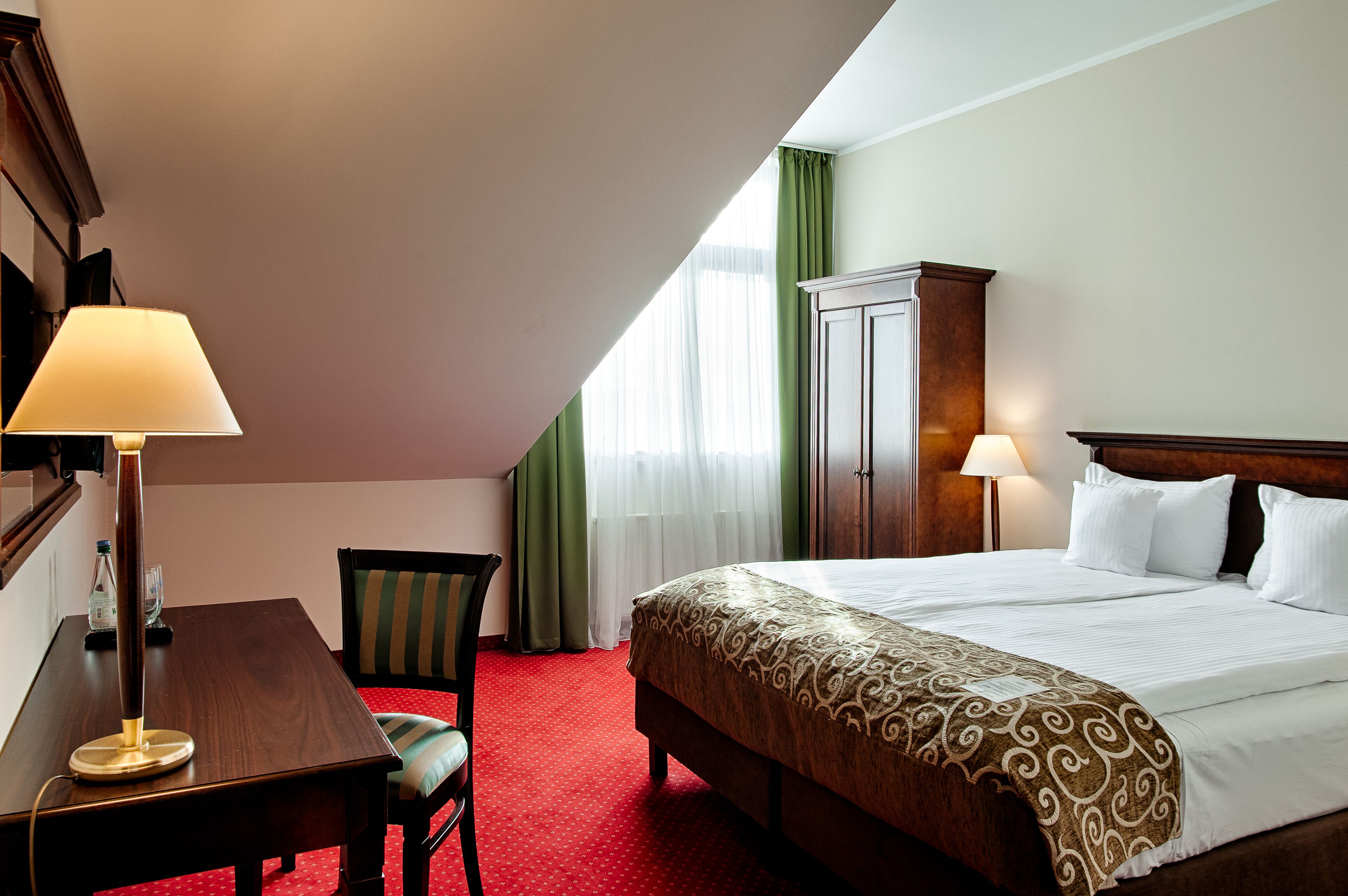 Una habitación de hotel con una cama, dos lámparas de noche, una mesita de noche de madera, una silla, un escritorio y un armario.