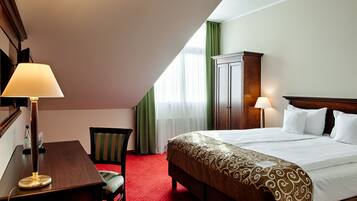 Una habitación de hotel con una cama, dos lámparas de noche, una mesita de noche de madera, una silla, un escritorio y un armario.