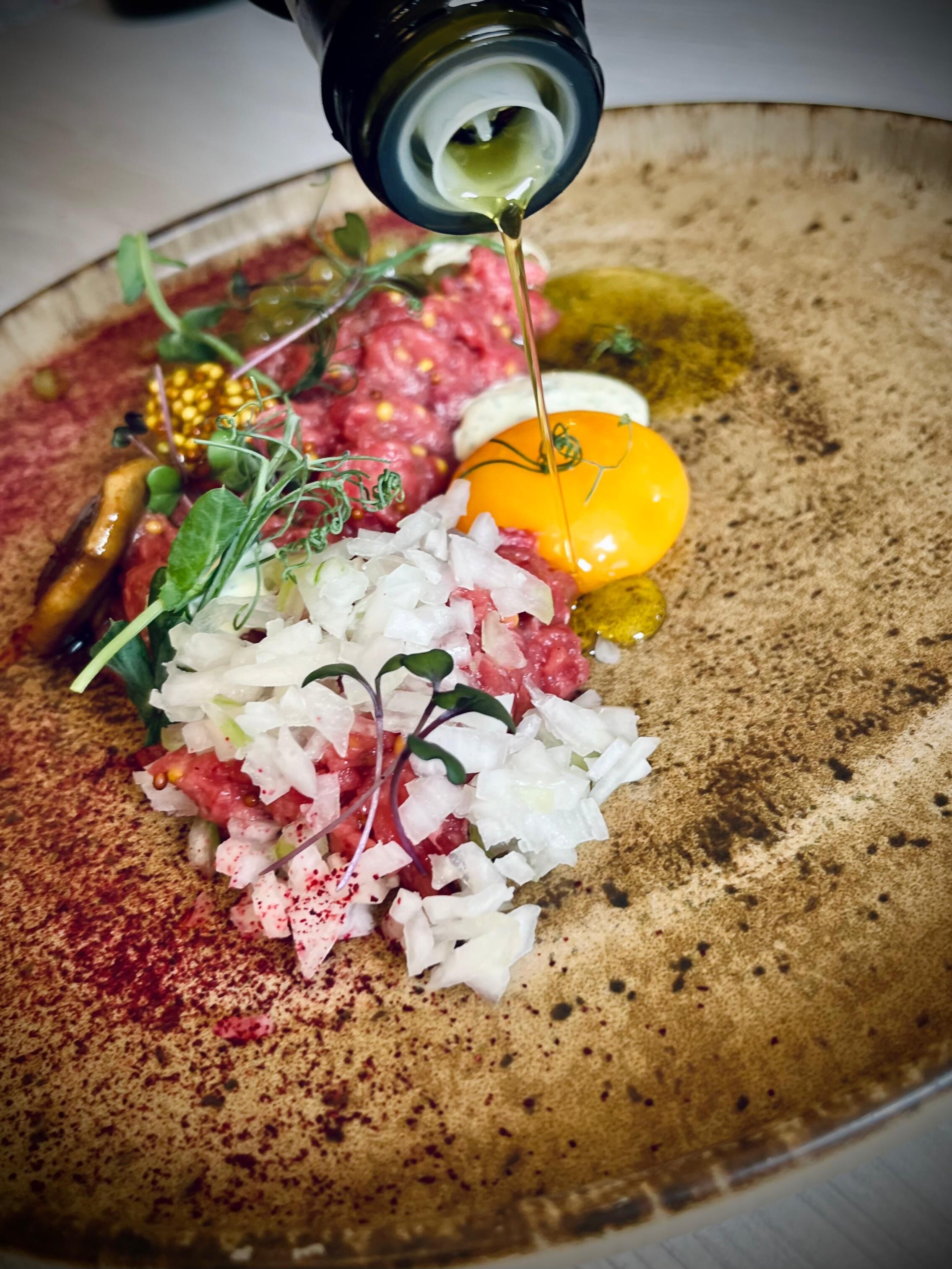 Ein Detailbild von einem Teller mit rohem Fleisch, Gemüse und einem gelben Eigelb, das mit Öl beträufelt wird.