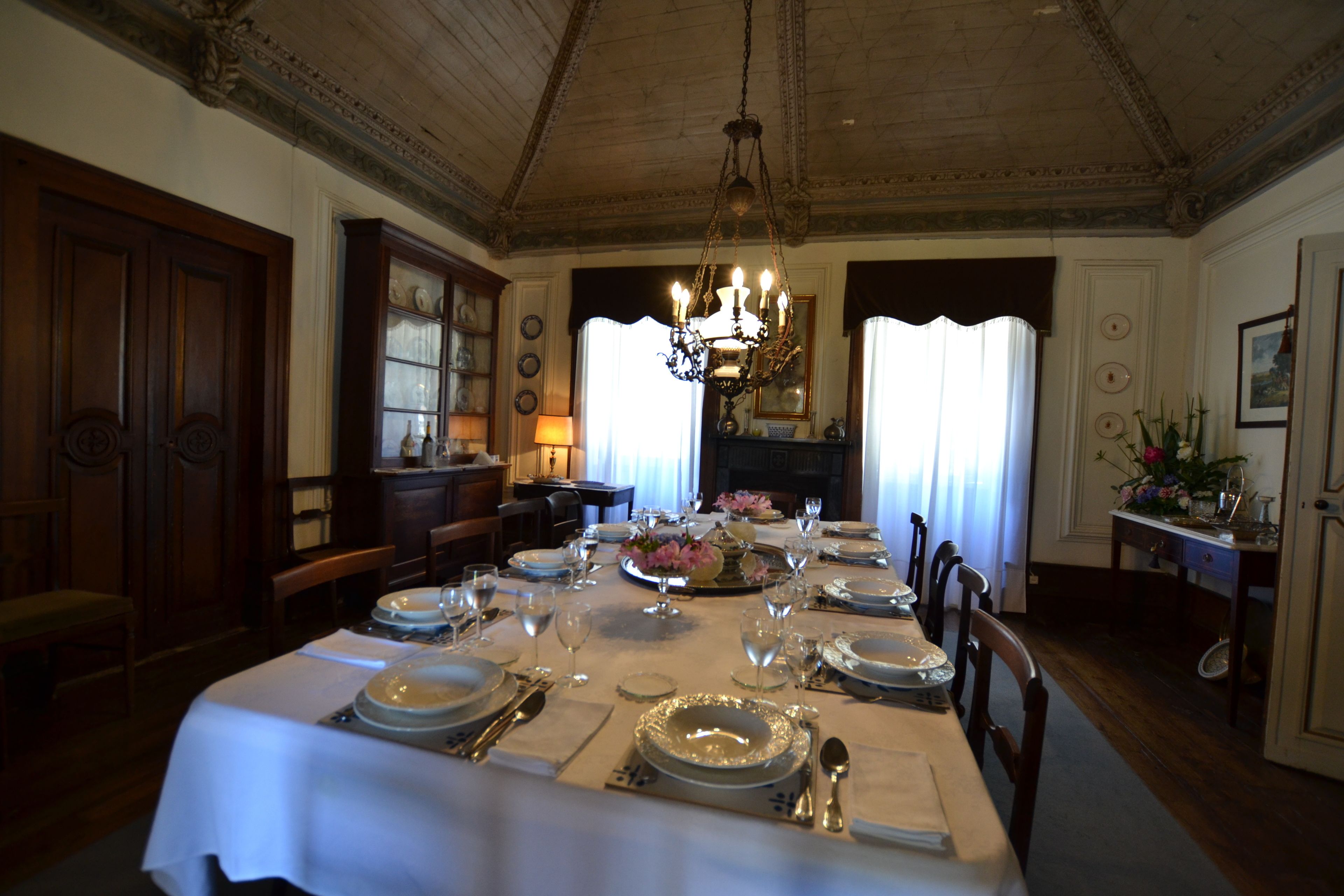 A formal dining room with a long table set for a meal, a chandelier, and a view of the dining area through a window.