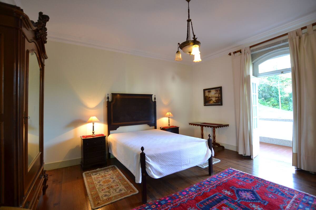 A bedroom with a large bed, two bedside tables, a nightstand, a wooden dresser, a chandelier, and a window with curtains.