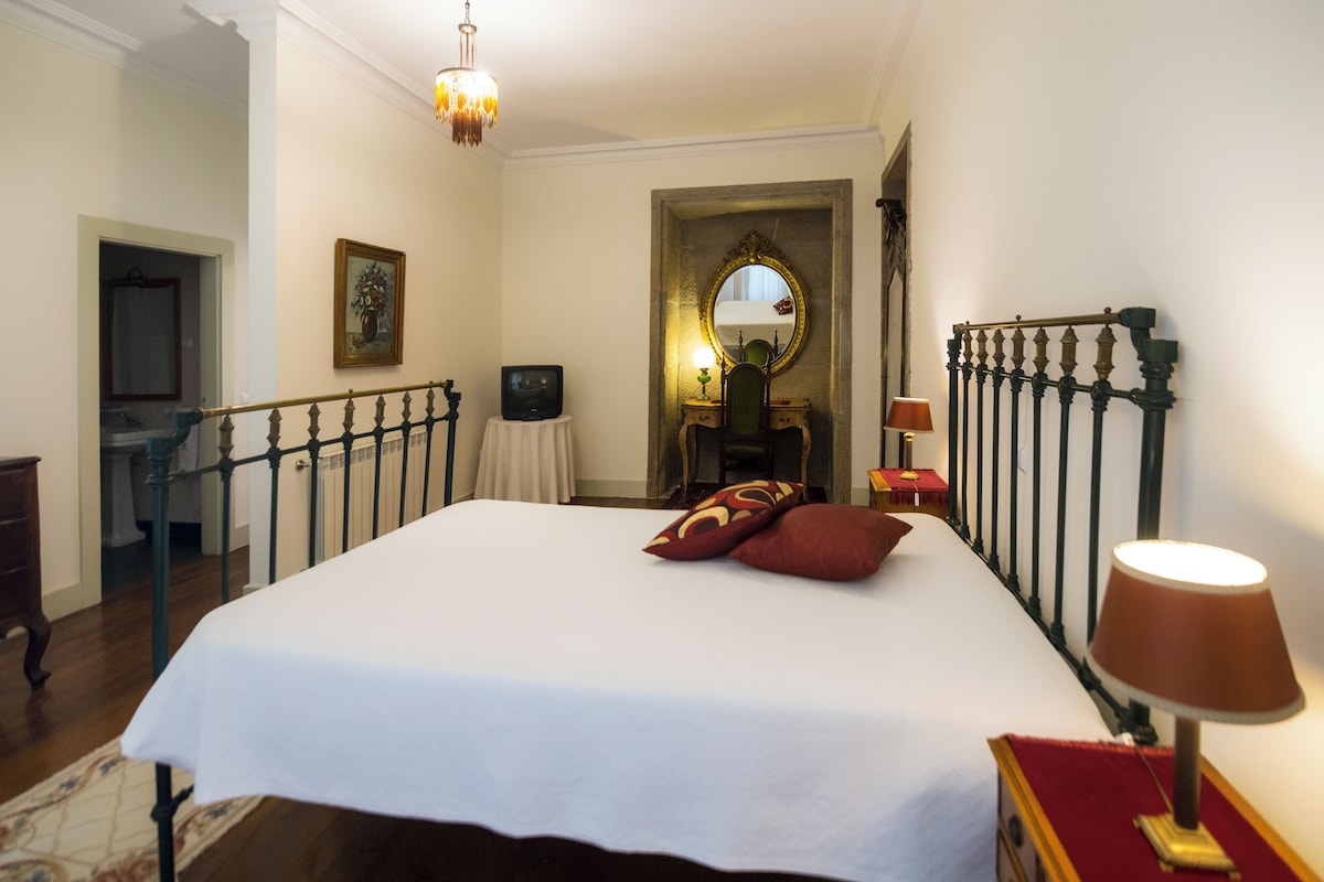 Hotellihuone, jossa on suuri sänky, työpöytä lampun kanssa, peili ja näkymä käytävälle.